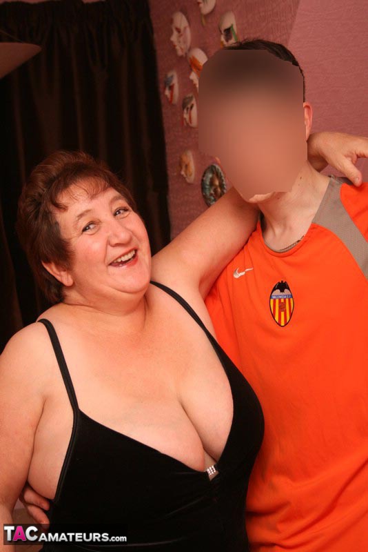 Hot granny teases a young stud with her chubby body in black shirt, white skirt, black stockings and white high heels then lets him suck her big breasts on a brown bed before she sucks his dick. - XXXonXXX - Pic 1