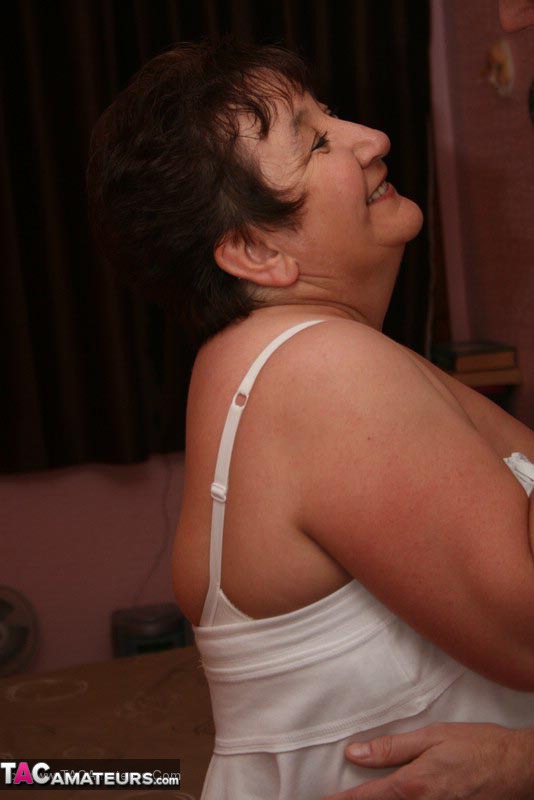 Mature BBW teases her horny hubby with her large body in white dress, lingerie, stockings and black high heels before she gets on her knees and wanks his cock on a brown bed. - XXXonXXX - Pic 9
