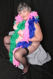 Hot mature babe displays her huge juggs as she pose naked wearing her black stockings and high heels while wrapped with multi-colored feather boas on a white chair.
