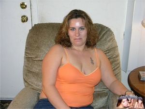 Chubby hottie sits on a gray sofa and displays her fat body before she slowly peels off her orange shirt, jeans and blue panty then bares her huge boobs and indulging pussy in different positions on a red bed. - Picture 1