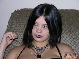 Goth babe takes off her black panty and teases with her indulging pussy in different positions on a gray couch wearing her black dress, bra, stockings and boots then gets naked and reveals her large breasts on a white bed. - Picture 4