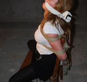 Busty brunette in white top and black pants roped tight to chair and gagged