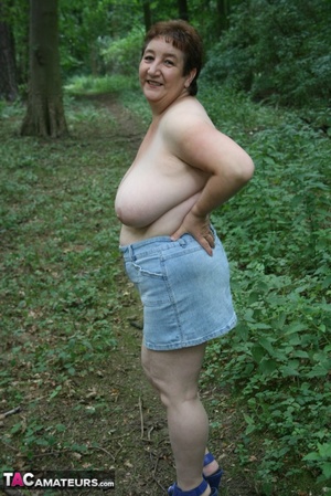 Chubby granny pose in the woods then peels off her black shirt and releases her giant tits before she pulls down her jeans skirt and teases with her humongous ass then pulls down her blue panty and expose her lusty pussy wearing her blue high heels. - Picture 5