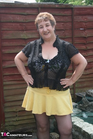 Steaming hot granny peels off her black blouse and yellow skirt and displays her chubby body in multi, colored underwear, black stockings and yellow high heels outdoor. - Picture 1