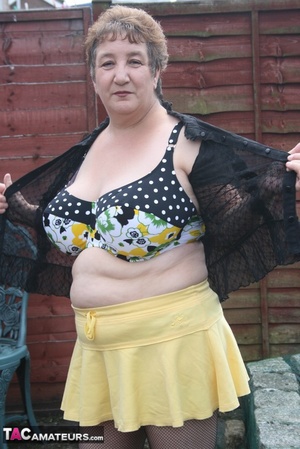 Steaming hot granny peels off her black blouse and yellow skirt and displays her chubby body in multi, colored underwear, black stockings and yellow high heels outdoor. - Picture 9