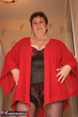 Chubby granny takes off her red robe and displays her her plus size body in red and black lingerie, black stockings and red high heels in different poses before she reveals her monster breasts on a green stairway. - XXXonXXX - Pic 1