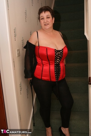 Cute granny peels off her black see through blouse and teases with her plus size body in different poses on a green stairway wearing her red and black corset, black leggings and high heels. - Picture 14