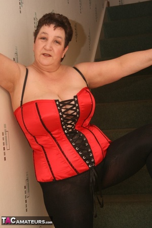 Cute granny peels off her black see through blouse and teases with her plus size body in different poses on a green stairway wearing her red and black corset, black leggings and high heels. - Picture 20