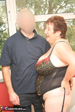 Plus size granny in black lingerie, stockings and high heels makes out with her hubby before she lets him suck her big breasts then rubs his dick between them before she eats his dick while she lets him rub her twat on a white bed. - Picture 1