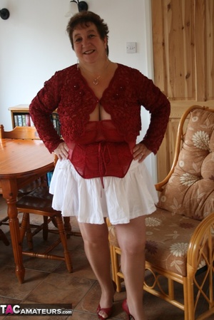 Chubby granny peels off her red blouse and white skirt and reveals her huge breasts before she pulls down her white panty and expose her lusty pussy wearing her red nighty, brown stockings and red high heels as she spreads wide on a brown couch. - Picture 1