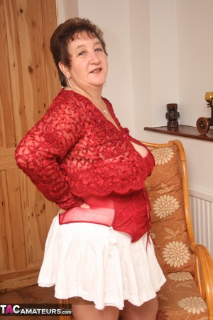 Chubby granny peels off her red blouse and white skirt and reveals her huge breasts before she pulls down her white panty and expose her lusty pussy wearing her red nighty, brown stockings and red high heels as she spreads wide on a brown couch. - Picture 2