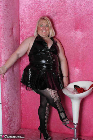 Big blonde in black leather outfit and black stockings stripteasing by the pink wall and showing her pierced vagina - Picture 1